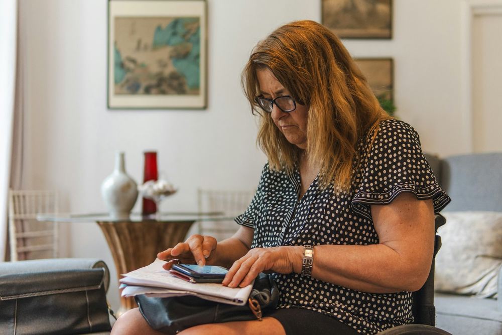 Eine Frau mit Brille sitzt im Wohnzimmer, schaut auf ihr Handy und hält Unterlagen auf dem Schoß.